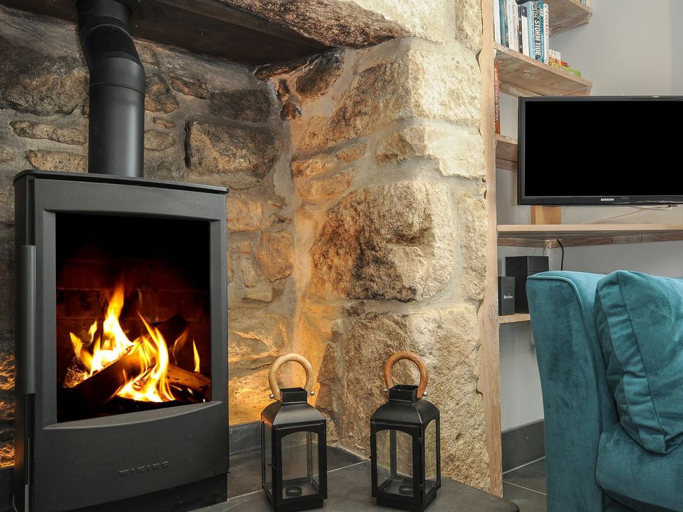 A wood burning stove with a fire inside stone walls two black lanterns a television and a blue chair at Stargazy Cottage in Praa Sands