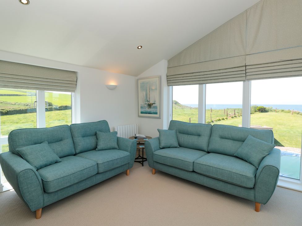 A living room with two teal sofas and large windows showing a grassy landscape and sea at 7 Ocean's Edge in Thurlestone