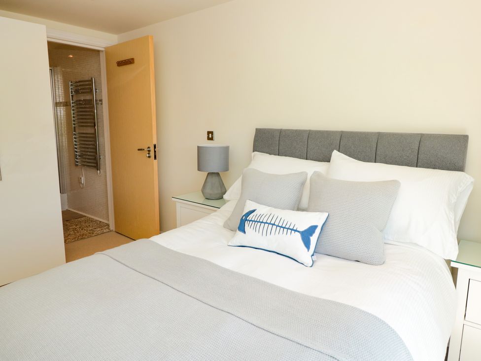 A bedroom with a bed with gray and white bedding a bedside table with a lamp and an open door leading to a bathroom at 7 Ocean's Edge in Thurlestone