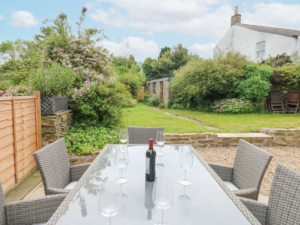 A garden patio with a glass table, six wicker chairs, wine bottle and glasses at Valley View in West Witton