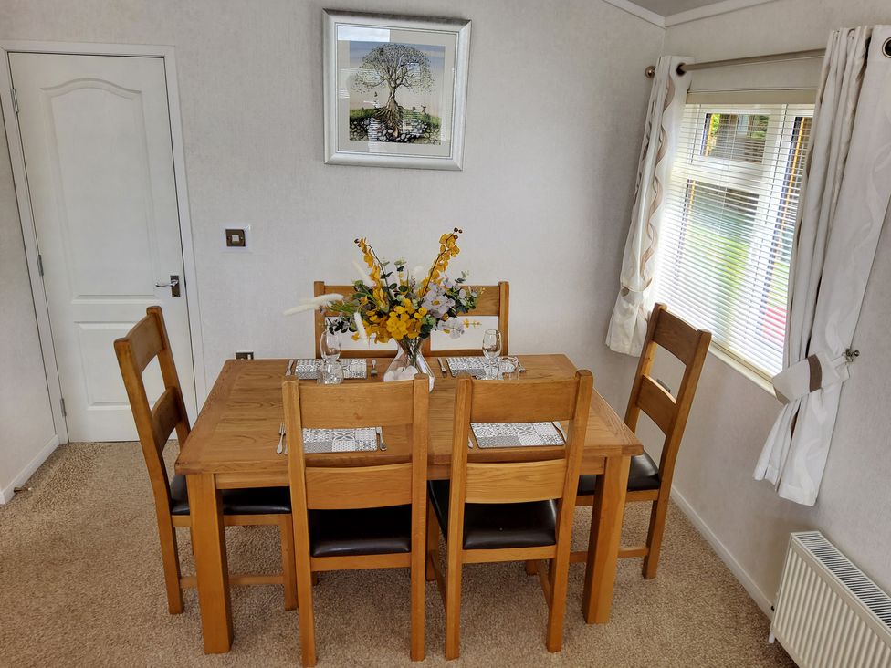 A dining room with a wooden table and four chairs with a flower vase on the table next to a window with curtains at Lodge 11 in Tavistock