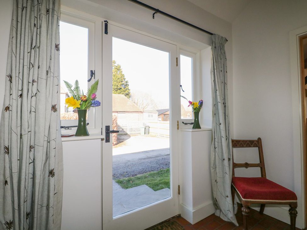 A glass door with floral curtains and vases of flowers on the windowsills next to a wooden chair with a red cushion at The Cowshed in Sedlescombe near Westfield