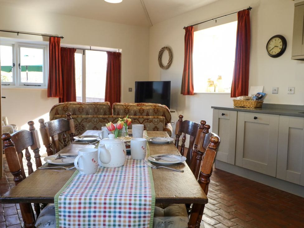 A dining table set with mugs, plates, and a teapot in a room with wooden chairs, a sofa, and kitchen cabinets at The Cowshed in Sedlescombe near Westfield