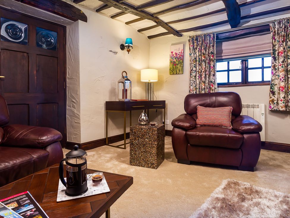 A living room with two leather armchairs a coffee table with a French press a side table with a lamp and a window with floral curtains at Tipsy Gin Cottage in Crosthwaite