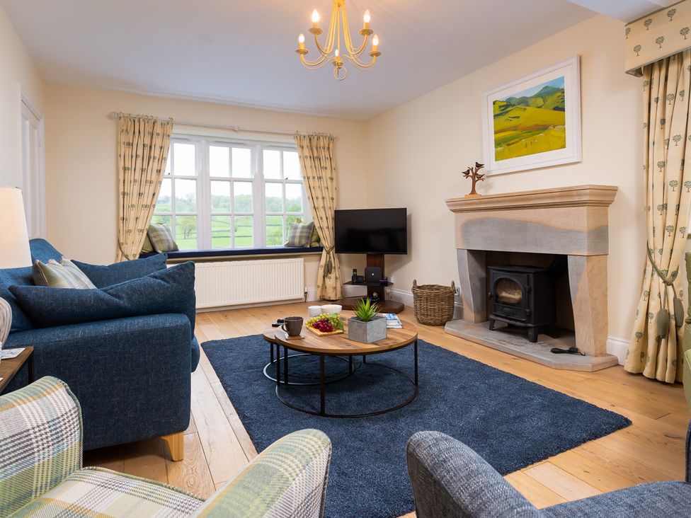 A living room with a blue sofa a wood stove a round wooden coffee table and a window with curtains at Grass Ings in Langdale