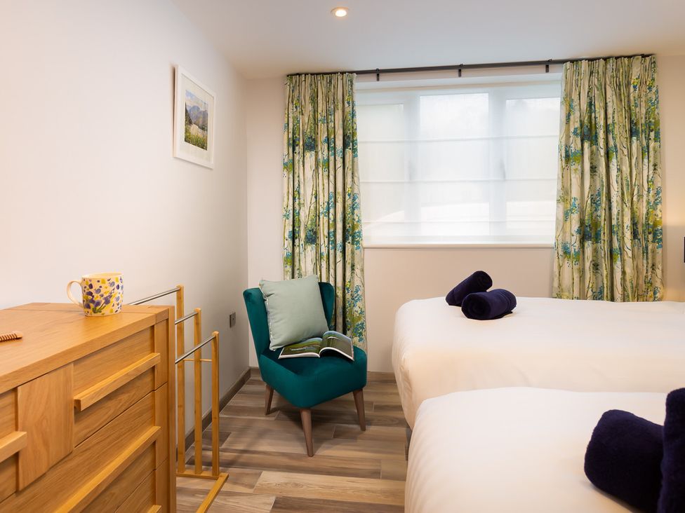 A bedroom with two beds with towels on them a green armchair with a pillow and book a wooden dresser a window with floral curtains at Fell View in Keswick