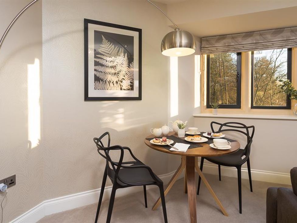 A small dining area with a round wooden table, two black chairs, a metal floor lamp, and a window showing trees outside at Hygge at No 8 in Windermere