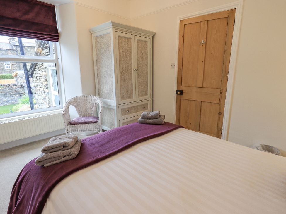 A bedroom with a bed with folded towels on a purple runner a wicker chair a white wardrobe and a wooden door at Ash Cottage in Windermere