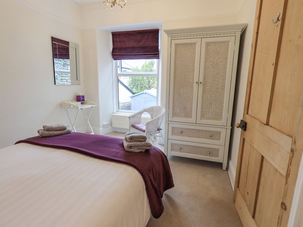A bedroom with a bed covered with white and purple linens a white wardrobe a white chair a small wall mirror and a wooden door at Ash Cottage in Windermere