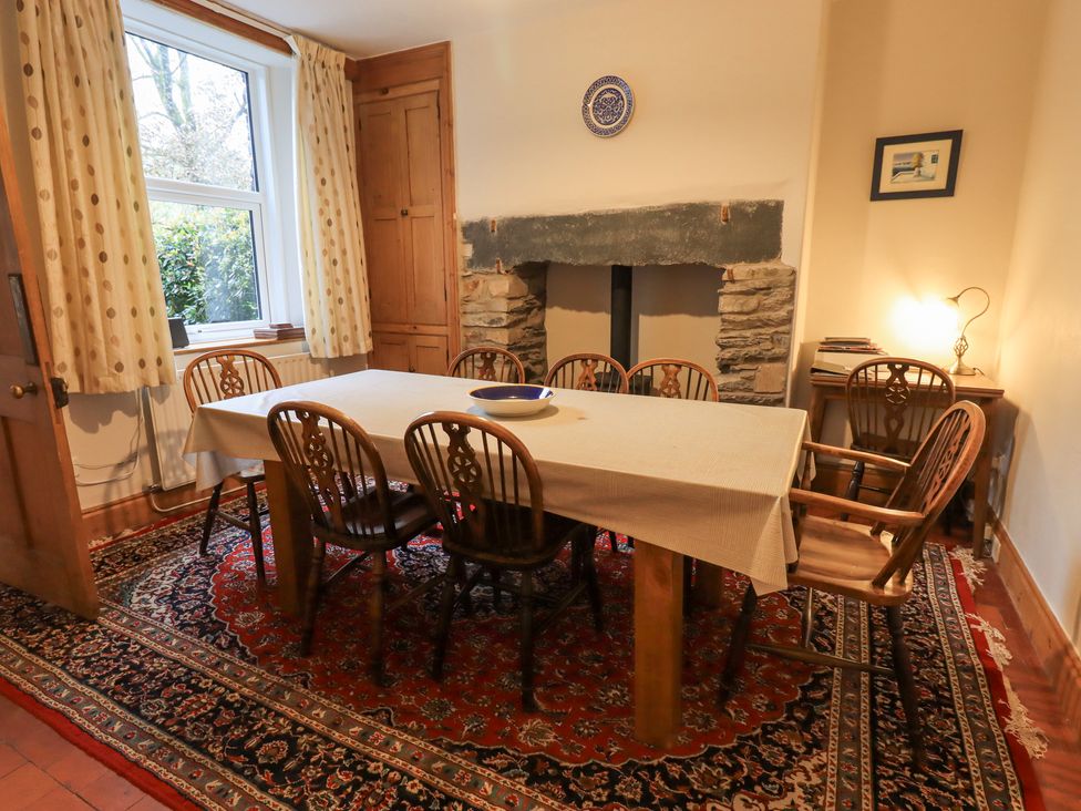 A dining room with a table covered by a cloth surrounded by wooden chairs and a fireplace with a stone surround at Millans Garth in Ambleside