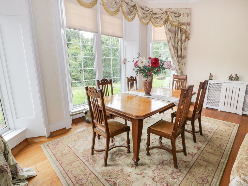 A dining room with wooden table and six chairs a vase with flowers on the table and large windows with curtains at Auchentroig House in Buchlyvie