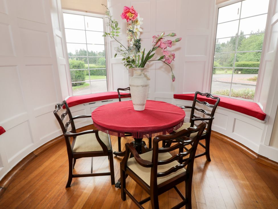 A round table with a red tablecloth and vase with flowers surrounded by four wooden chairs in a room with large windows and red cushioned window seats at Auchentroig House in Buchlyvie
