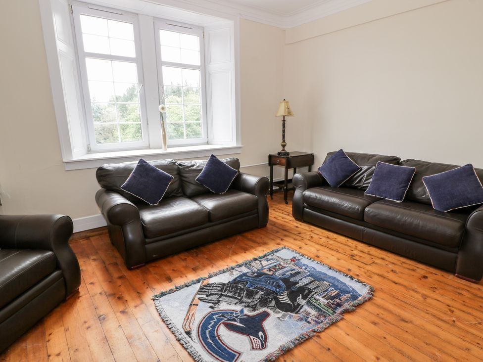 A living room with two black leather sofas with blue cushions a wooden floor a small side table with a lamp and a window with a single flower at Auchentroig House in Buchlyvie