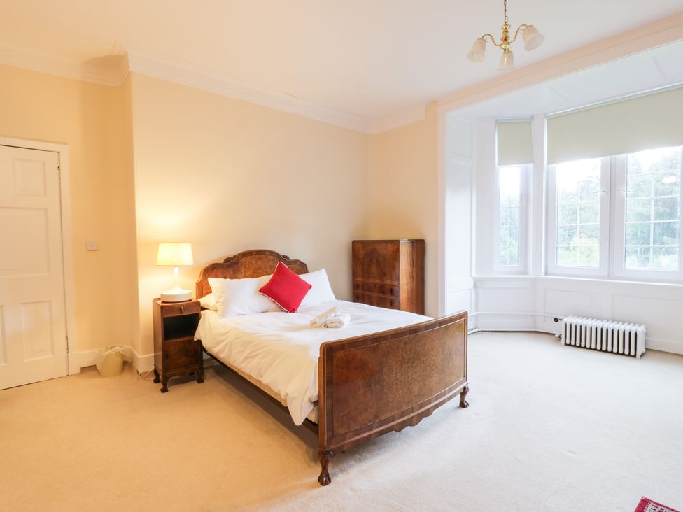A bedroom with a wooden bed with white bedding and a red pillow a wooden nightstand with a lamp a chest of drawers a white radiator and large windows at Auchentroig House in Buchlyvie