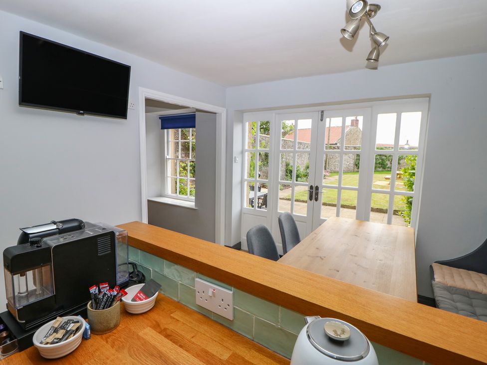A kitchen counter with a coffee machine and condiments overlooking a dining table and chairs with glass doors leading to a garden at Hall Cottage in Ovington