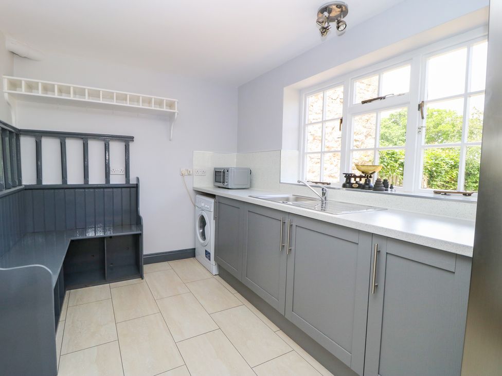 A kitchen with gray cabinets a washing machine a microwave a bench and large windows showing greenery at Hall Cottage in Ovington