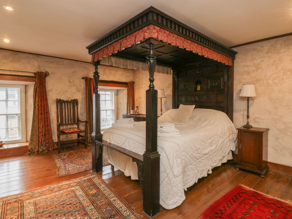 A bedroom with a four poster bed two windows with curtains an armchair two rugs and a bedside table with a lamp at The Pend in Whithorn
