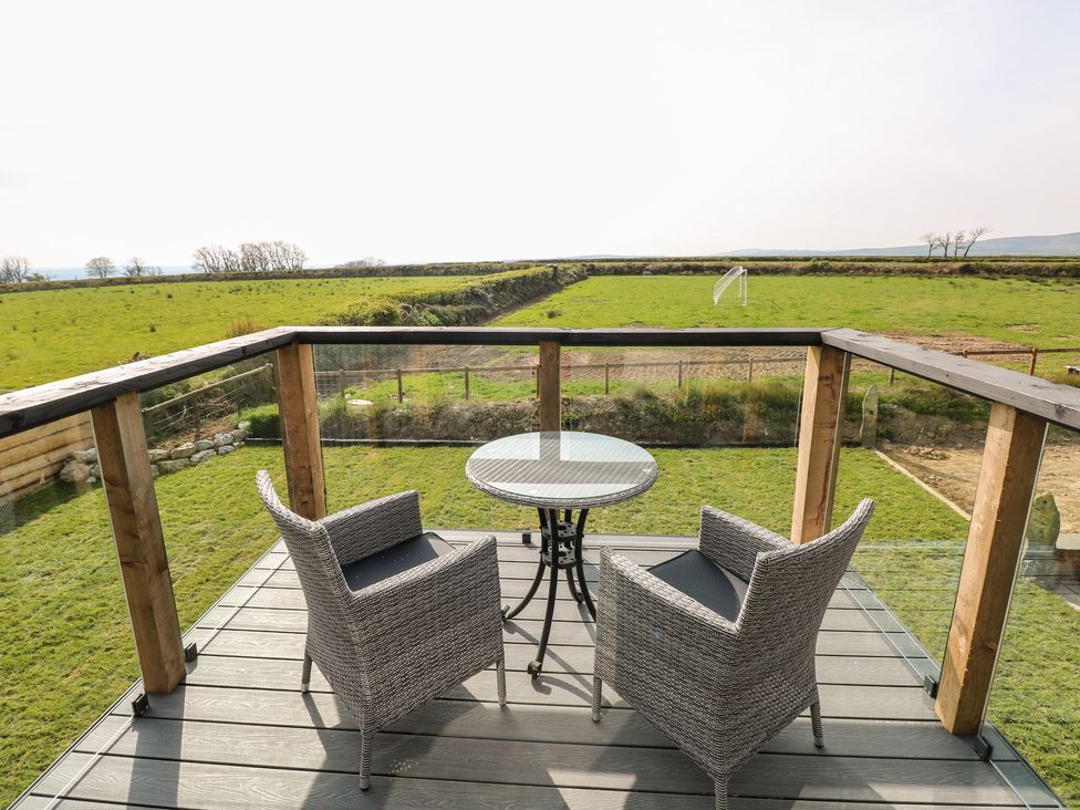 A balcony with two wicker chairs and a round glass table overlooking a grassy field at Glowty Brynbedw in Efailwen