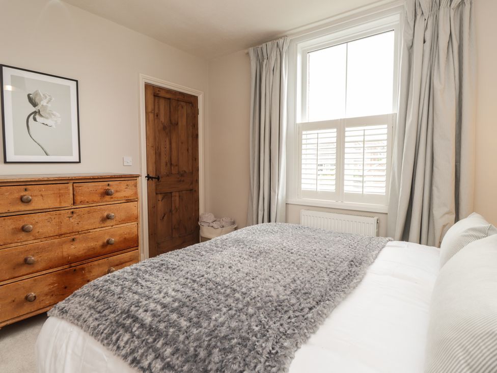 A bedroom with a wooden chest of drawers a wooden door a window with curtains and a bed with a gray blanket and white pillows at Vine Cottage in Tenterden