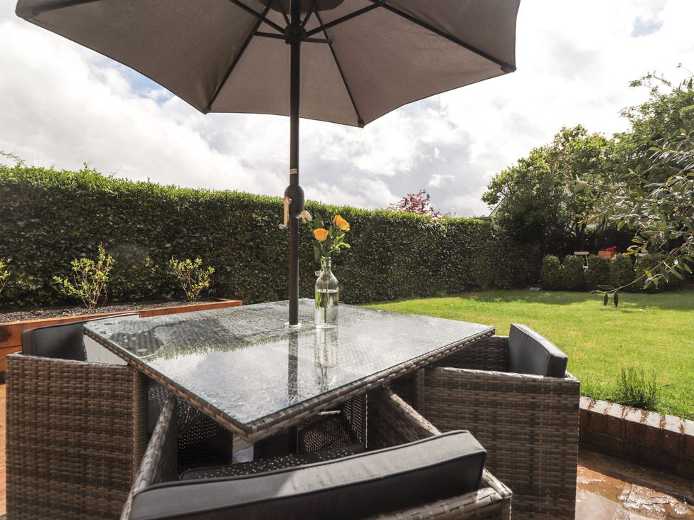 An outdoor patio table with glass top and an umbrella surrounded by wicker chairs with cushions in a garden at Vine Cottage in Tenterden