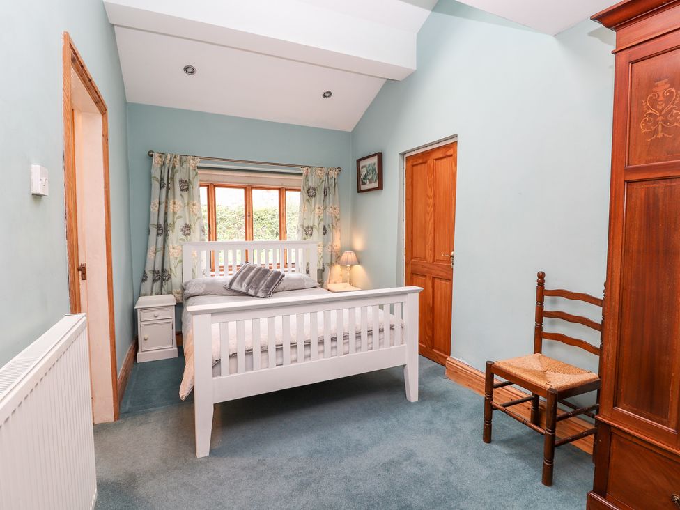 A bedroom with a white bed frame two bedside tables a wooden chair a wooden wardrobe and a window with floral curtains at Abbey Cottage in Denbigh
