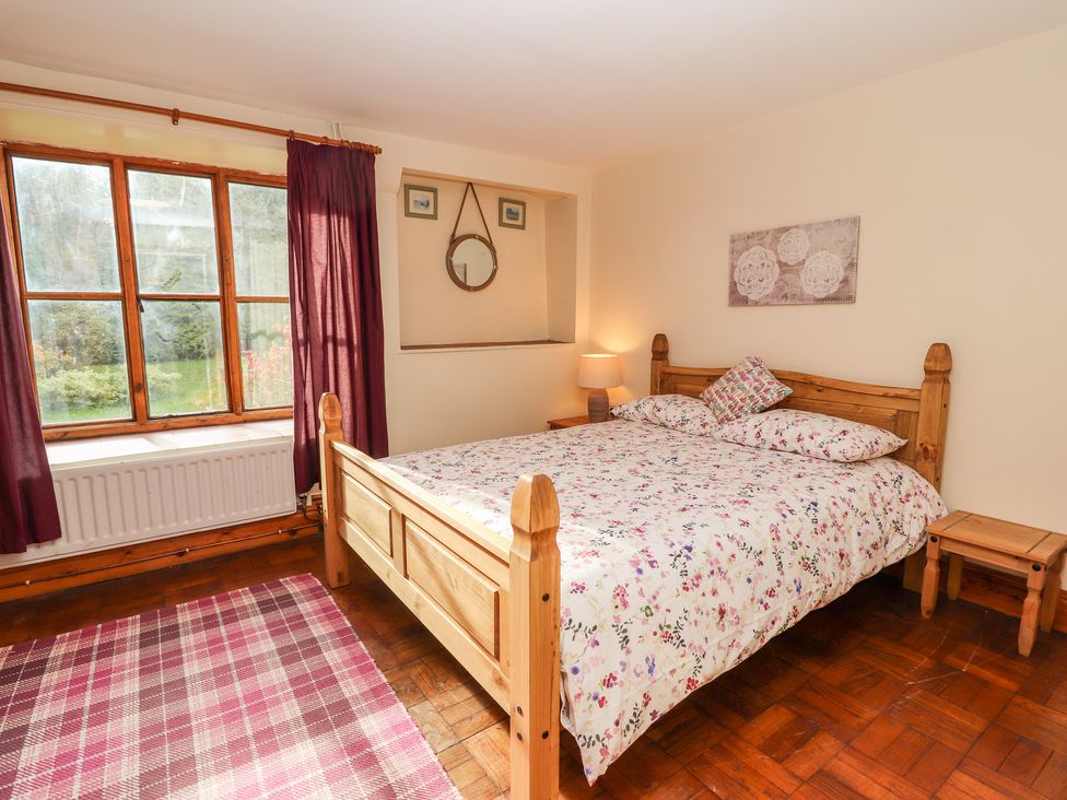 A bedroom with a wooden bed, floral bedding, a window with curtains, a rug, and bedside tables at Abbey Cottage in Denbigh