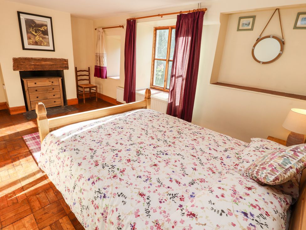 A bedroom with a wooden bed with floral bedding curtains on the windows a wooden chair and a chest of drawers in the fireplace at Abbey Cottage in Denbigh