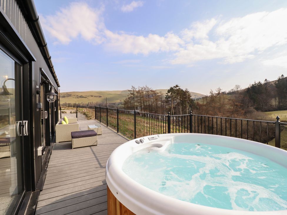 A deck with a hot tub sofa and table overlooking hills and trees at The Sheep Shed in Llanrhaeadr-Ym-Mochnant