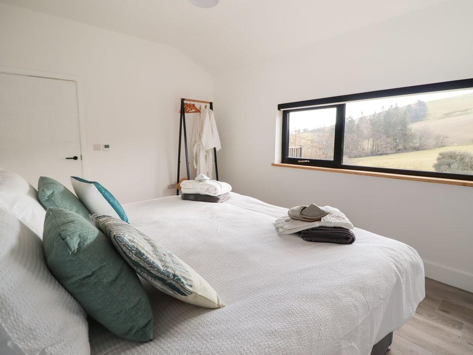 A bedroom with a white bedspread pillows and folded towels on the bed a window showing a countryside view and a clothes rack with robes at The Sheep Shed in Llanrhaeadr-Ym-Mochnant