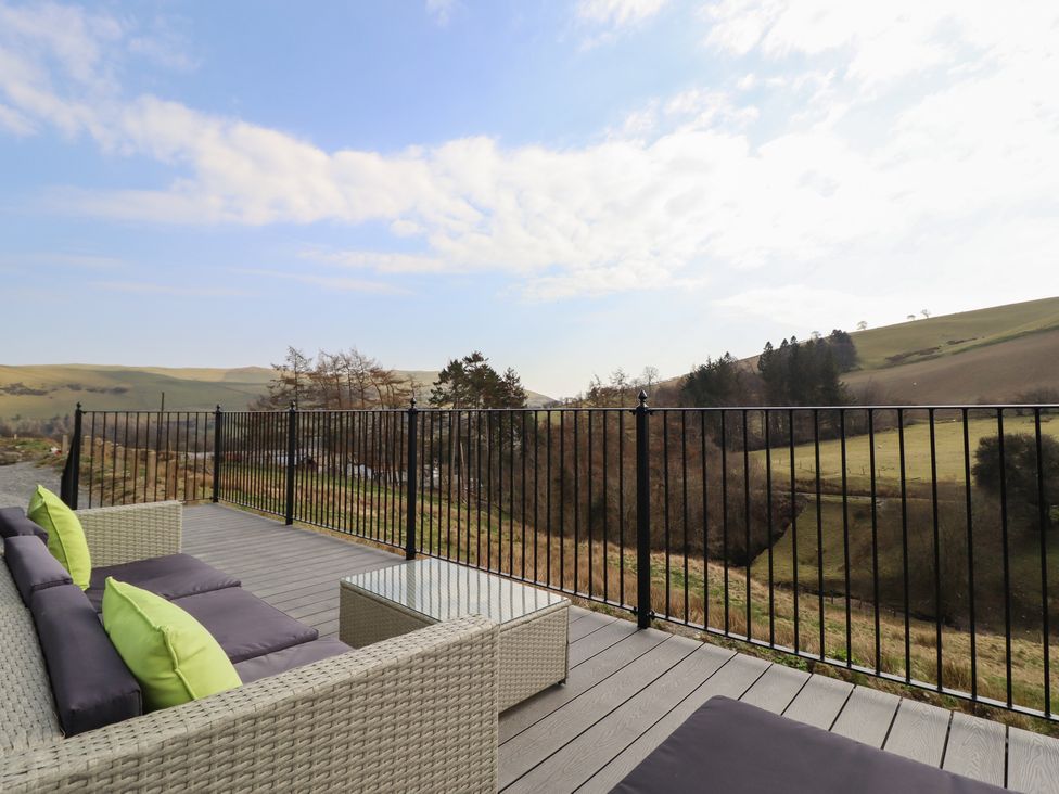 An outdoor deck with wicker sofa seating cushions and a glass coffee table overlooking a grassy hillside at The Sheep Shed in Llanrhaeadr-Ym-Mochnant