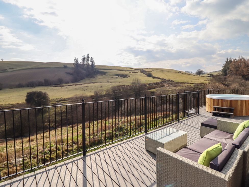 An outdoor deck with patio furniture a glass table and a hot tub overlooking grassy hills and trees at The Sheep Shed in Llanrhaeadr-Ym-Mochnant