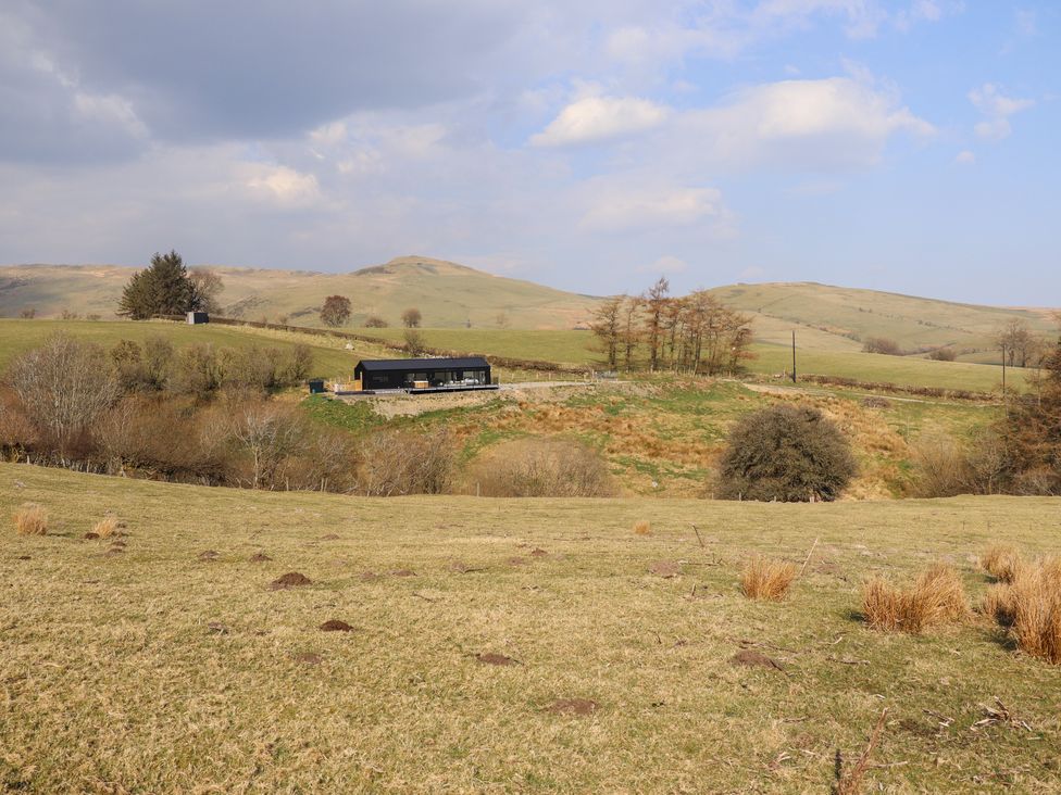 A grassy landscape with a black building and some trees on rolling hills at The Sheep Shed in Llanrhaeadr-Ym-Mochnant