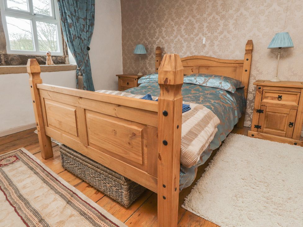 A bedroom with a wooden bed frame blue patterned bedding wooden nightstands with blue lamps and a window with blue curtains at Cobblers Cottage in Haworth
