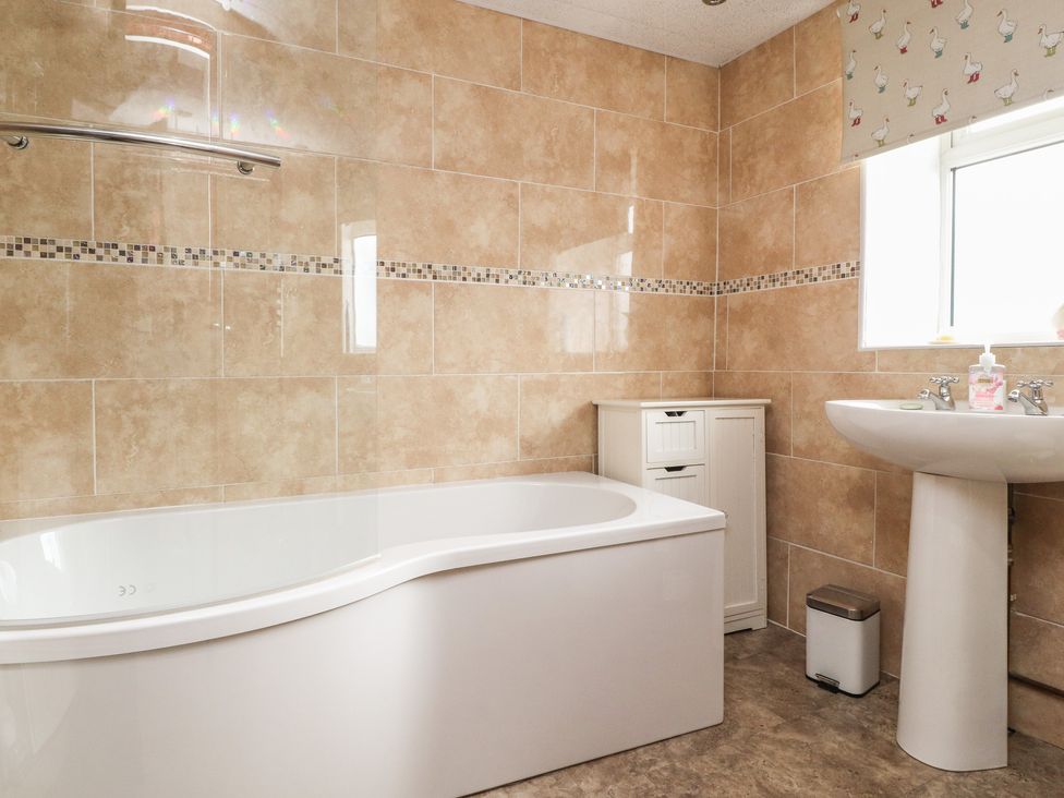 A bathroom with a white bathtub and a small cabinet next to a white pedestal sink with a window above at Cobblers Cottage in Haworth