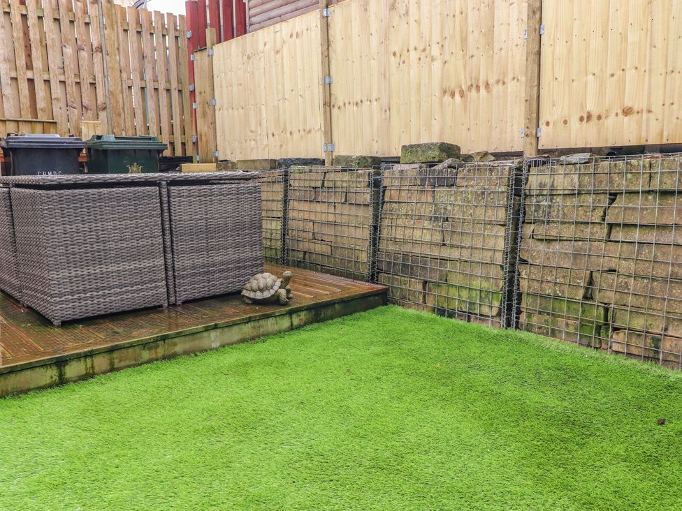An outdoor area with artificial grass a wooden deck with wicker furniture a stone turtle ornament and a stone wall with wooden fencing