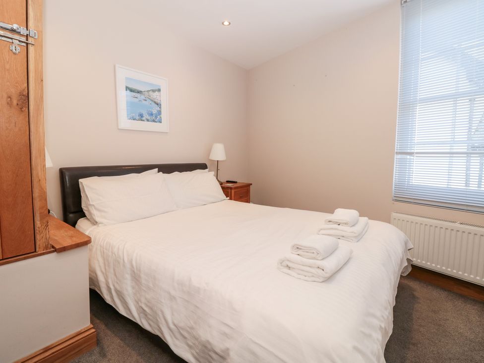 A bedroom with a double bed with white linens and folded towels on top a bedside table with a lamp window with blinds and a framed picture on the wall at Simples in Dartmouth
