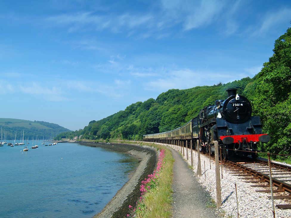 A steam train running on railway tracks next to a body of water with boats trees and hills at Simples in Dartmouth