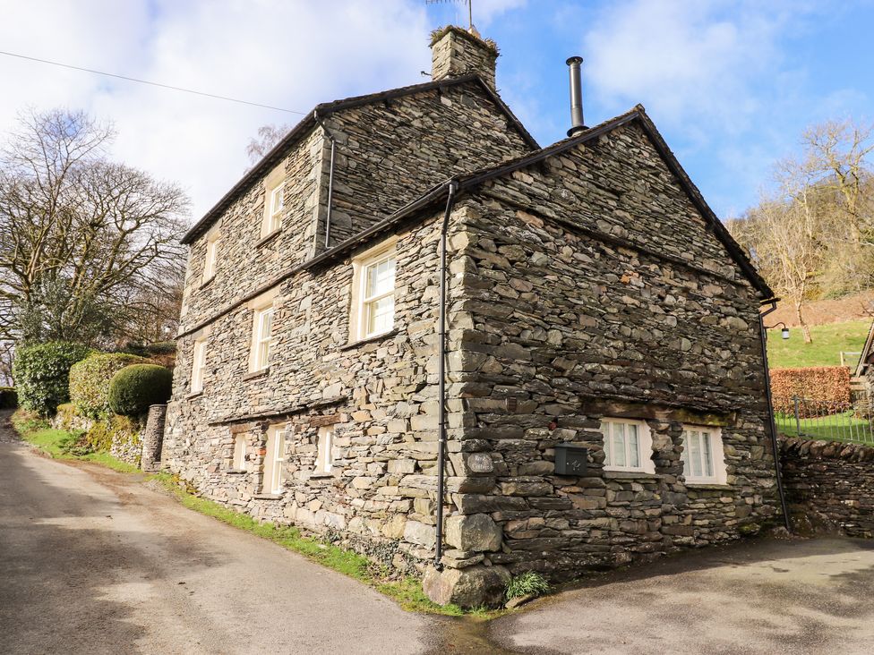 A stone cottage with multiple windows beside a road and trees at Rose Cottage At Troutbeck in Troutbeck
