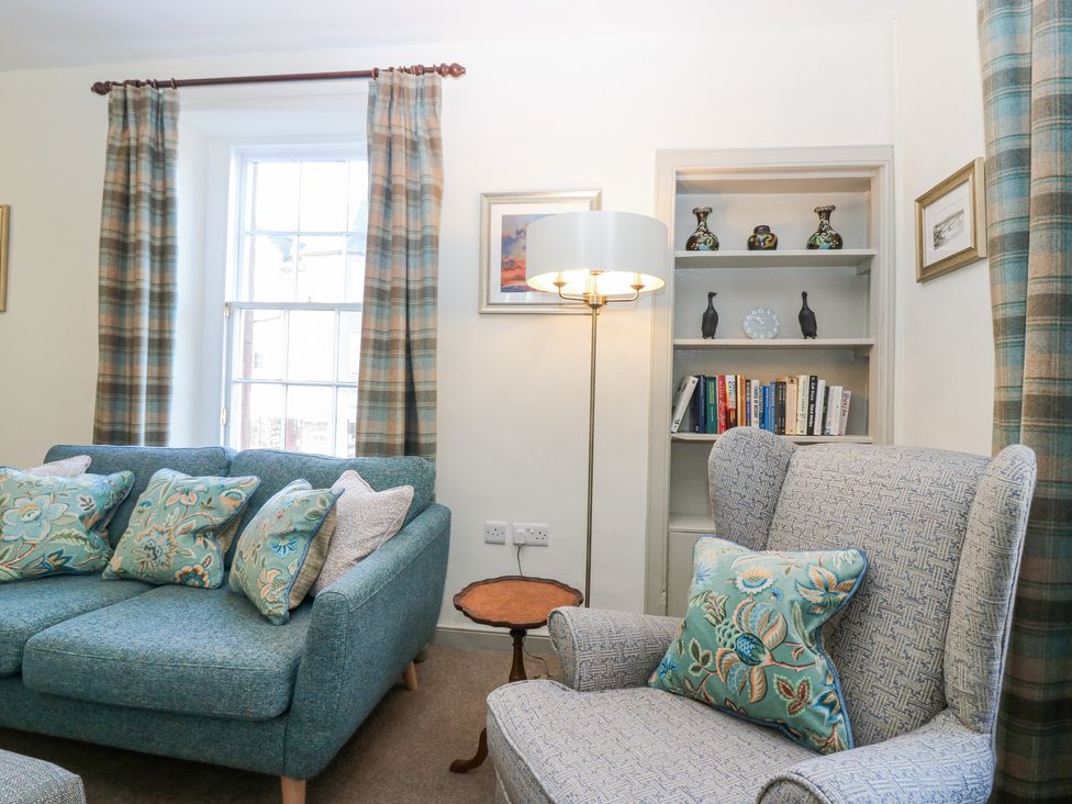 A living room with blue sofa floral cushions armchair plaid curtains and bookshelf at Pear Cottage - Priorwood Garden in Melrose
