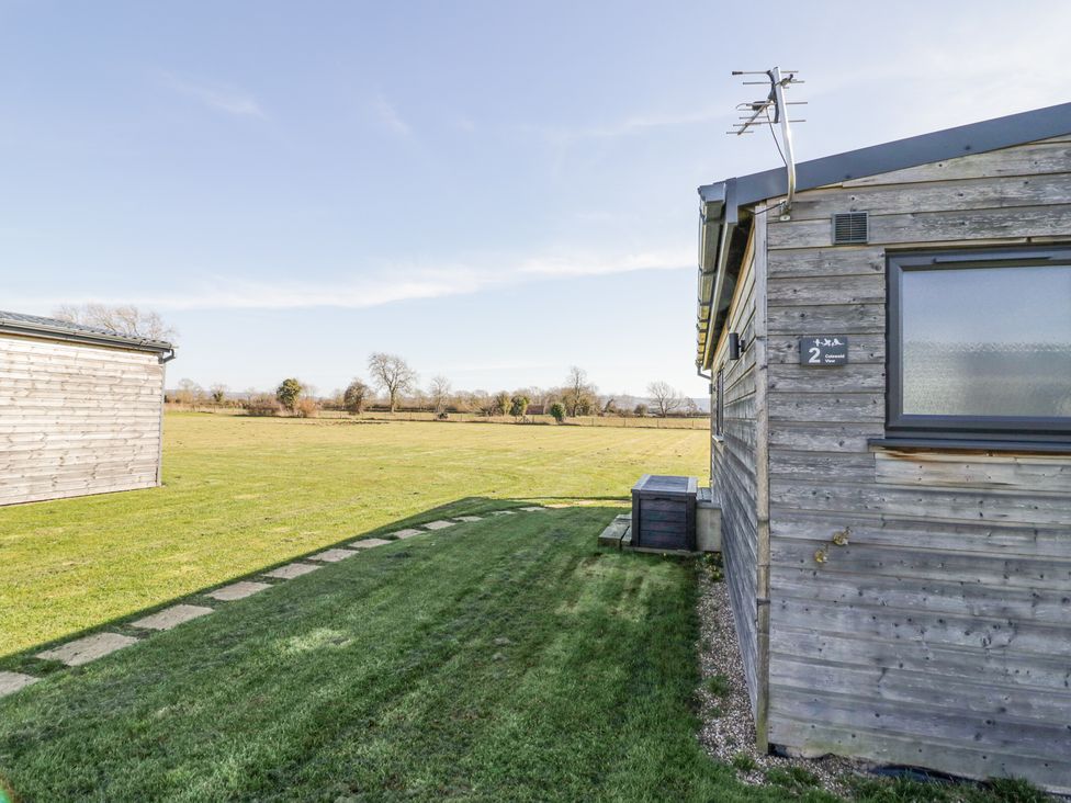An outdoor view showing a side of a wooden building with a small shed and a large grassy field at Cotswold View 2 in Honeybourne