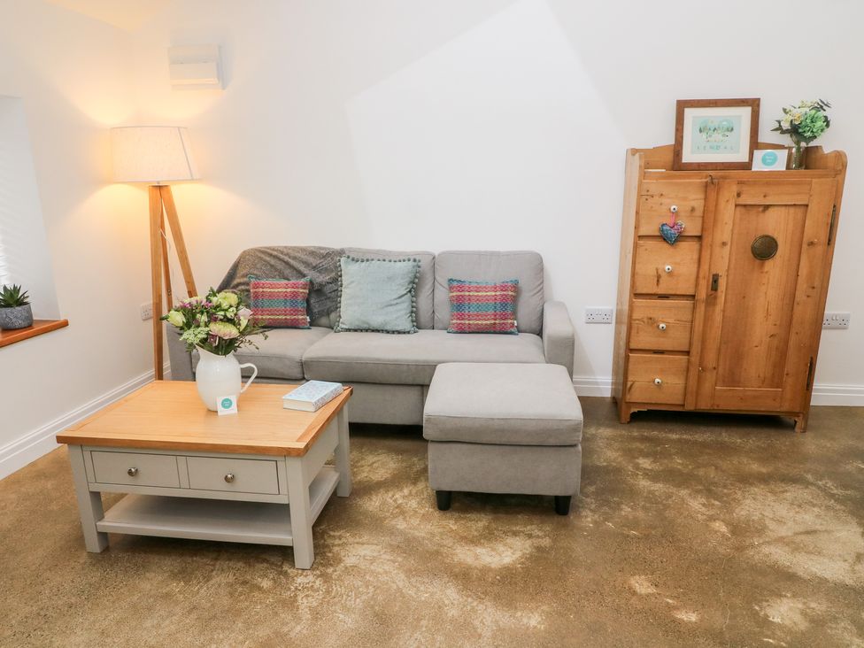 A living room with a gray sofa and ottoman a wooden coffee table with flowers and a book a wooden cabinet with decorations and a floor lamp at Horncop Nook Kendal