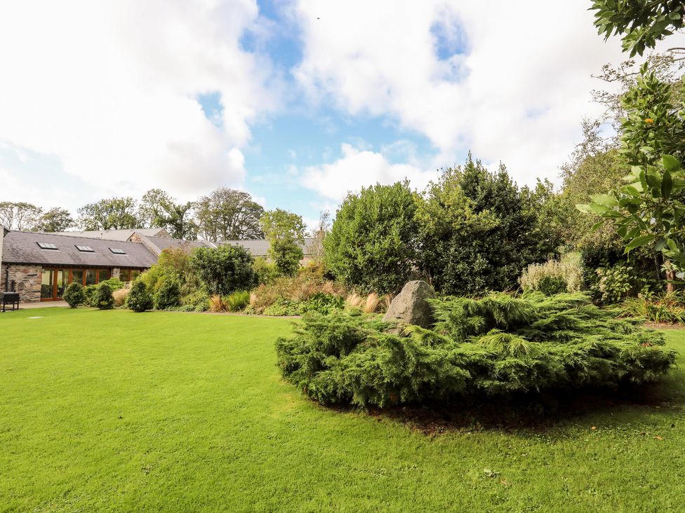 A grassy garden with bushes and a stone in the center next to a stone building with a slate roof at Clynnog House in Dwyran near Newborough