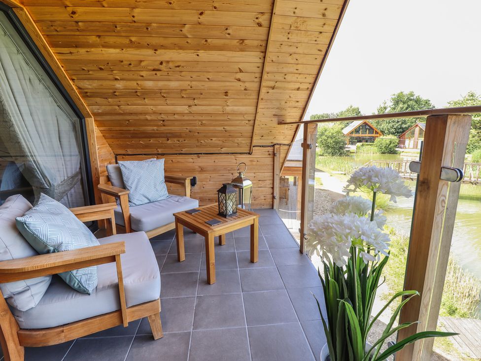 A balcony with wooden chairs cushions a small table lanterns and white flowers overlooking a pond at Lake View Lodge in Thorpe-On-The-Hill