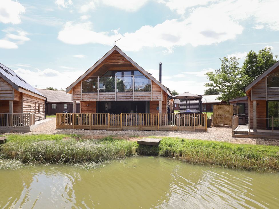 A wooden house with a balcony and a fenced patio next to a pond at Lake View Lodge in Thorpe-On-The-Hill