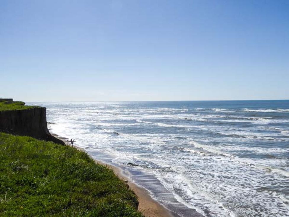 A coastal cliff with green grass overlooking a sandy beach and ocean waves under a clear sky at Chapel House in Chale