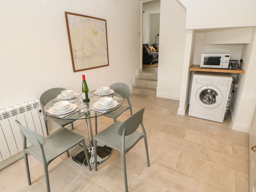 A dining area with a glass table set for four with dishes and a bottle a washing machine and microwave under a counter and stairs leading to a living room at The Old Store 2 Alpine Terrace in Reeth