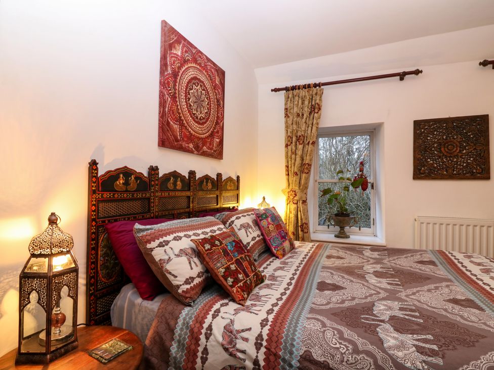 A bedroom with a bed featuring patterned bedding and pillows a decorative headboard lamp on a wooden table a window with curtains and a potted plant on the sill and wall art at Wonderland Cottage in Newburgh Fife