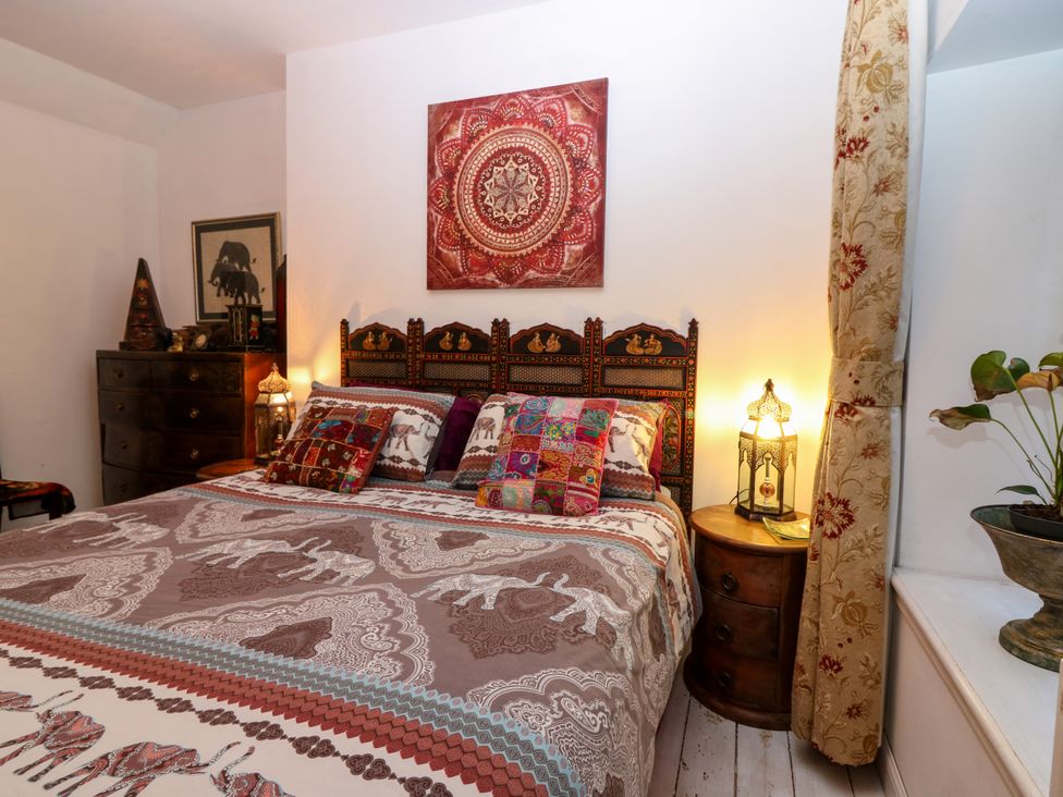 A bedroom with a patterned bedspread and pillows next to a wooden dresser and a round bedside table with a lamp at Wonderland Cottage in Newburgh Fife