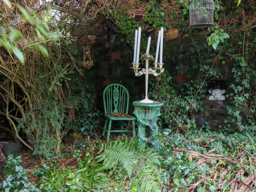 A green wooden chair and a green decorative pedestal with a white candelabra among dense foliage and ivy at Wonderland Cottage in Newburgh Fife