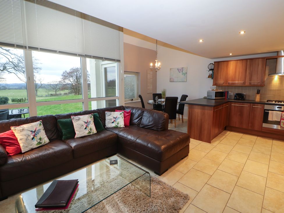 An open plan living area with a brown leather sectional sofa glass coffee table dining table and chairs and a kitchen with wooden cabinets at Lark Meadow in Kirkby Lonsdale